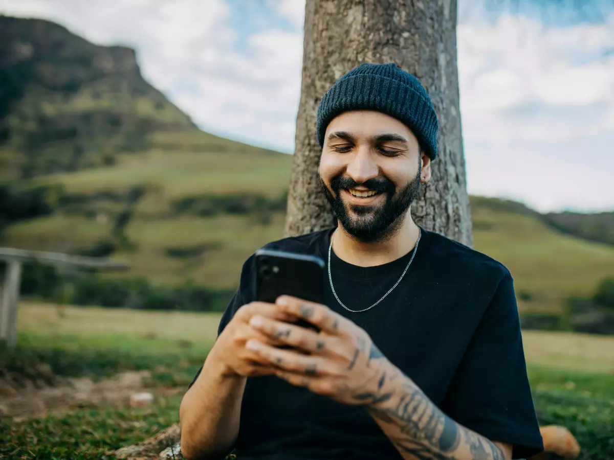 A young man with a beard and a black beanie is smiling while looking at his phone. He is standing in front of a tree with a green hill and blue sky in the background.